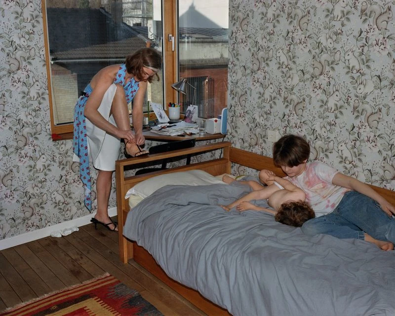 Woman in blue dress putting on shoes near bed with two children in a bedroom with floral wallpaper, bathed in natural light.