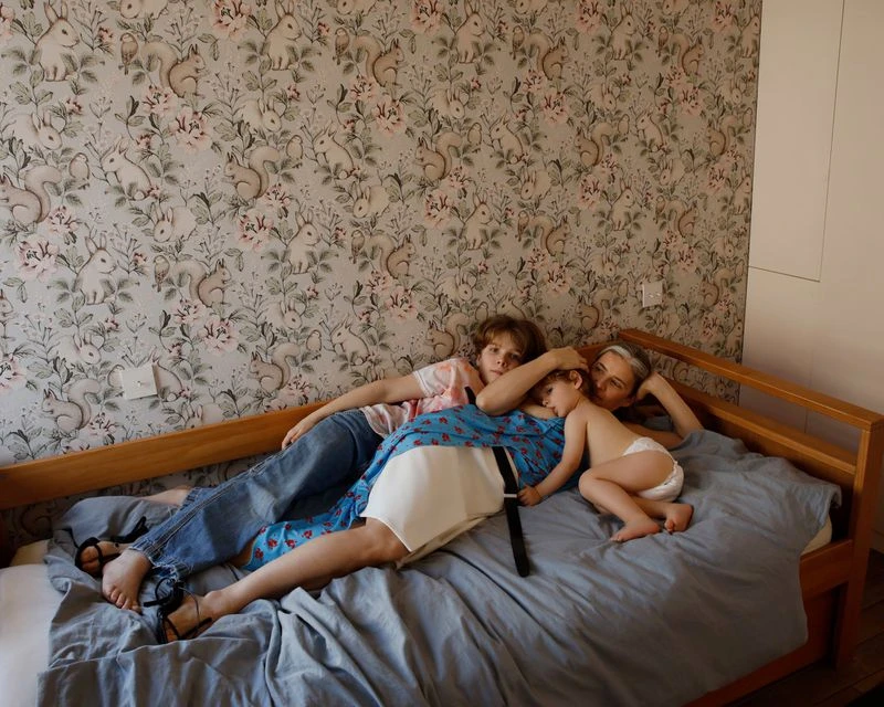 Family portrait on a bed featuring an adult woman, a young boy, and a toddler, indoors with patterned wallpaper and natural light.