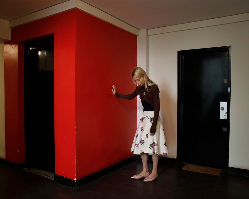 Woman in a floral skirt leans against a red wall in a hallway next to a black elevator door. Brown cardigan and blonde hair.