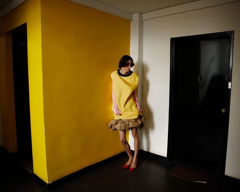 Adult female fashion model posing in yellow dress and red shoes, leaning on a yellow wall next to a black door in an indoor setting.