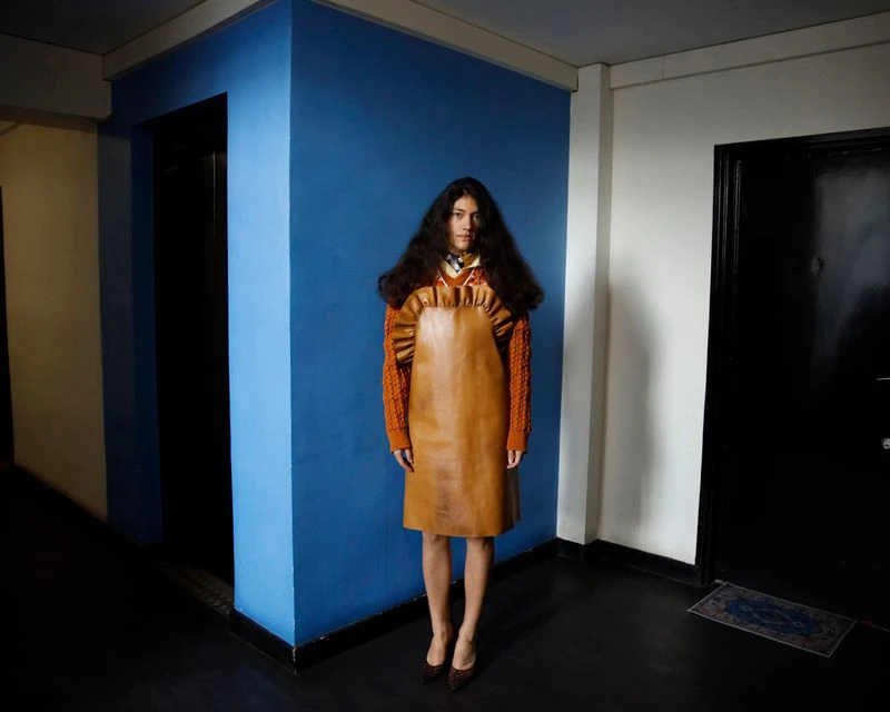Woman with long dark hair stands in a blue and white hallway wearing a tan leather dress, orange sweater, and dark shoes, in a fashion shot.