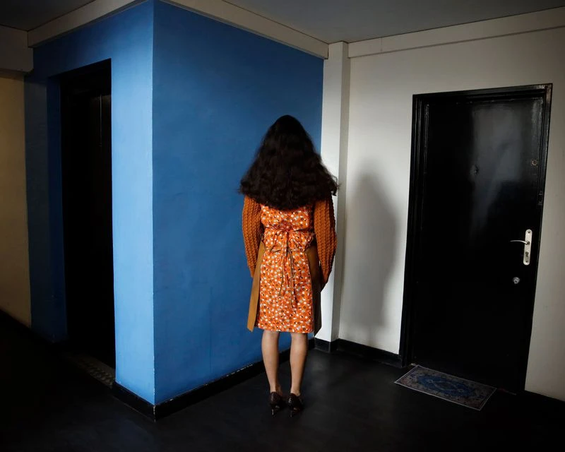 Woman with long dark hair stands in a hallway wearing an orange floral dress, an orange sweater, and black high heels.