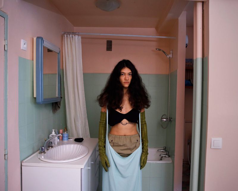 A woman in a black top and light blue skirt stands in a bathroom with green tile and a white sink, creating a striking fashion portrait.