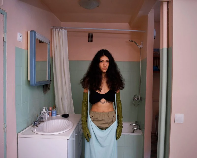 A woman in a black top and light blue skirt stands in a bathroom with green tile and a white sink, creating a striking fashion portrait.