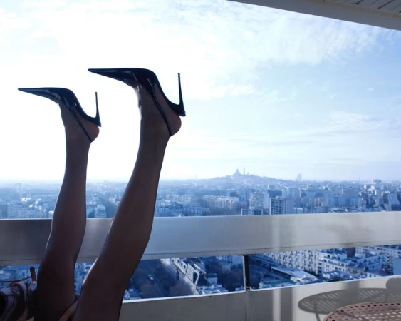 Legs of a woman in black pantyhose and high heels are raised up in the air on a balcony overlooking a cityscape and blue sky.