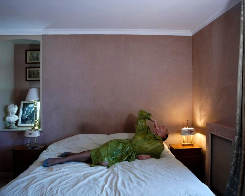 Adult female in green dress lies on white bed in indoor bedroom interior with pale-brown walls, two lamps, and white bust statue.