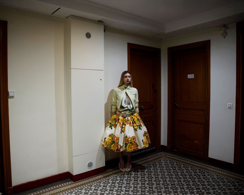 Fashionable woman in a vintage floral skirt and cropped jacket stands in a hallway with tiled floors and paneled doors.