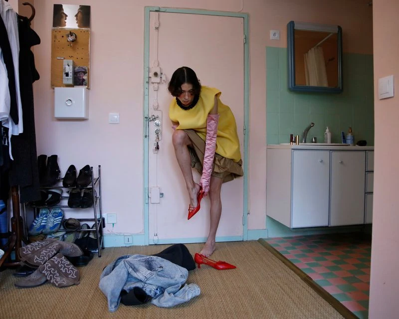 Fashionable person is indoors putting on a red high-heeled shoe in a domestic scene with various clothing and accessories scattered around.