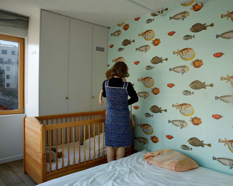A woman holds a toddler in a room decorated with fish wallpaper, featuring a crib, bed, and a window showing a building exterior.