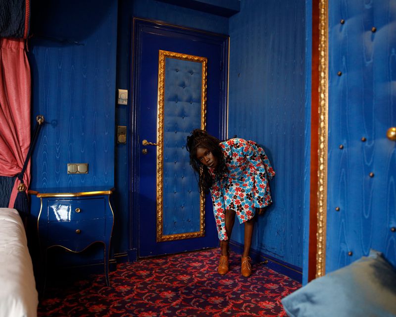 Elegant woman in a floral dress bending in a blue hotel room with golden accents, captured in a medium shot with soft lighting.