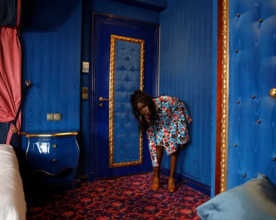 Elegant woman in a floral dress bending in a blue hotel room with golden accents, captured in a medium shot with soft lighting.