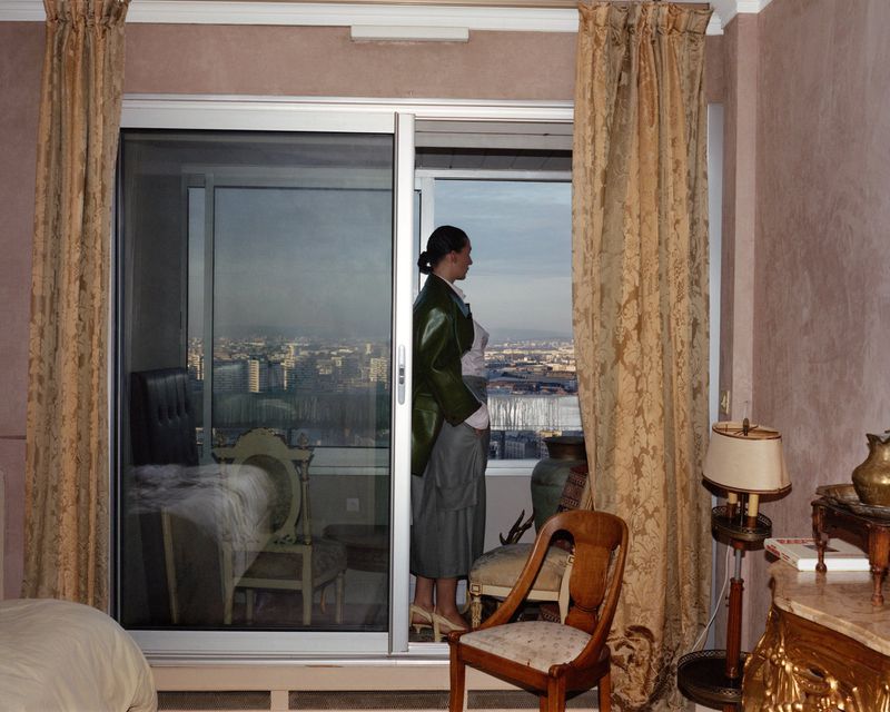 Woman in green jacket gazes out sliding glass door at city view. Draped curtains and bedroom furniture visible inside the room.