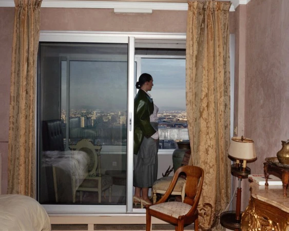 Woman in green jacket gazes out sliding glass door at city view. Draped curtains and bedroom furniture visible inside the room.