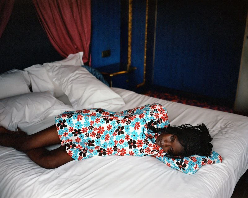 Young adult woman with dark skin and braided hair wearing a floral dress lying on a bed with white sheets and pillows in an indoor setting.
