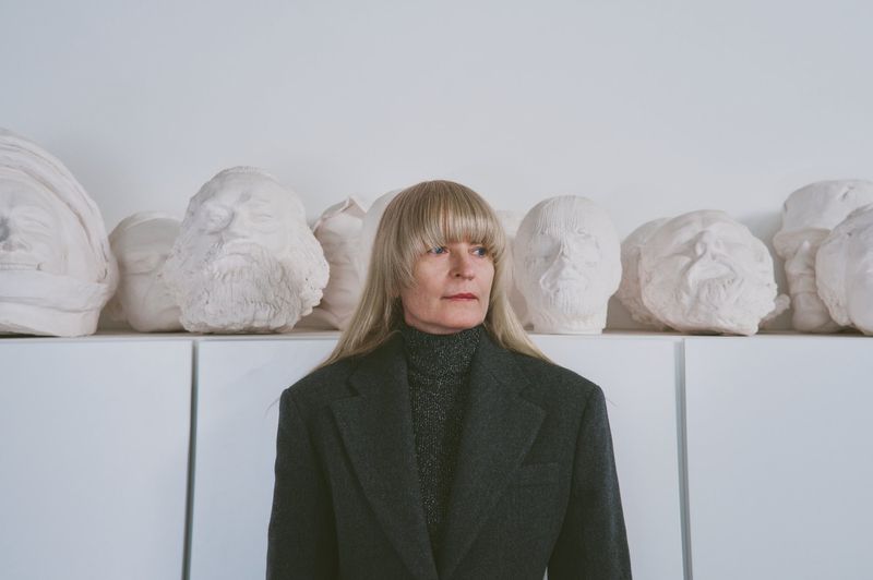 Portrait of an adult female wearing a coat, standing indoors with artful plaster head sculptures displayed on white shelving behind her.