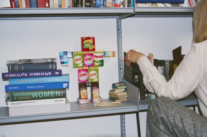 Conceptual art shelves: books stacked beside a figure constructed from laundry detergent boxes on a shelf, adult human reaches for book.