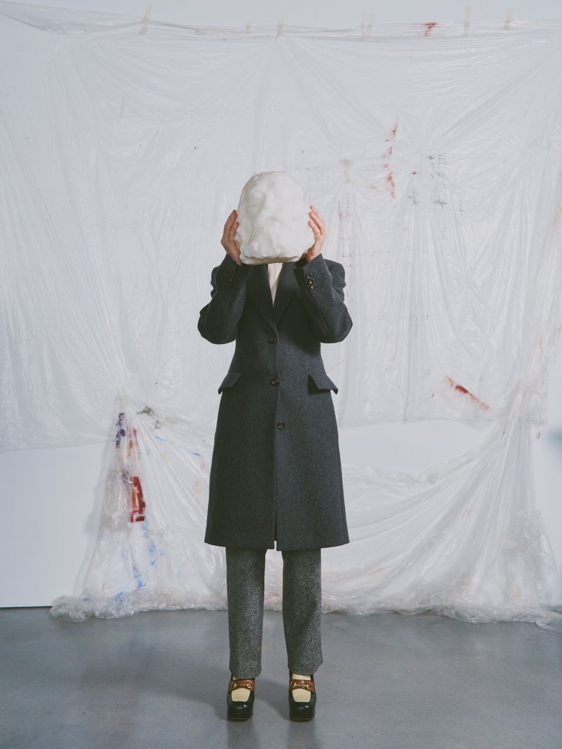 Person in overcoat holds white sculpted mask over face against a white backdrop with paint splatters in a studio setting.