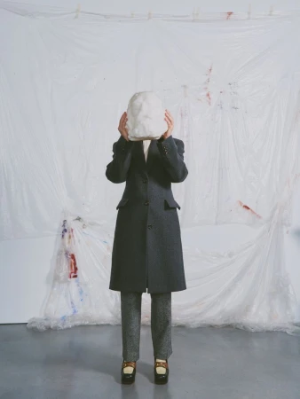 Person in overcoat holds white sculpted mask over face against a white backdrop with paint splatters in a studio setting.
