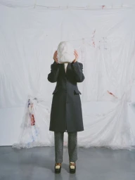 A person in a tailored charcoal coat holding a white sculpted bust over their face in front of a draped plastic backdrop.