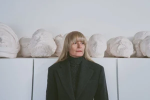 Artist Goshka Macuga in a charcoal wool blazer standing in front of a row of white plaster sculpted heads.