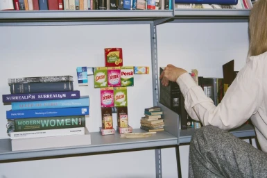 A person organizing a grey shelf beside art book stacks and a small robot figure constructed from colorful packaged product boxes.