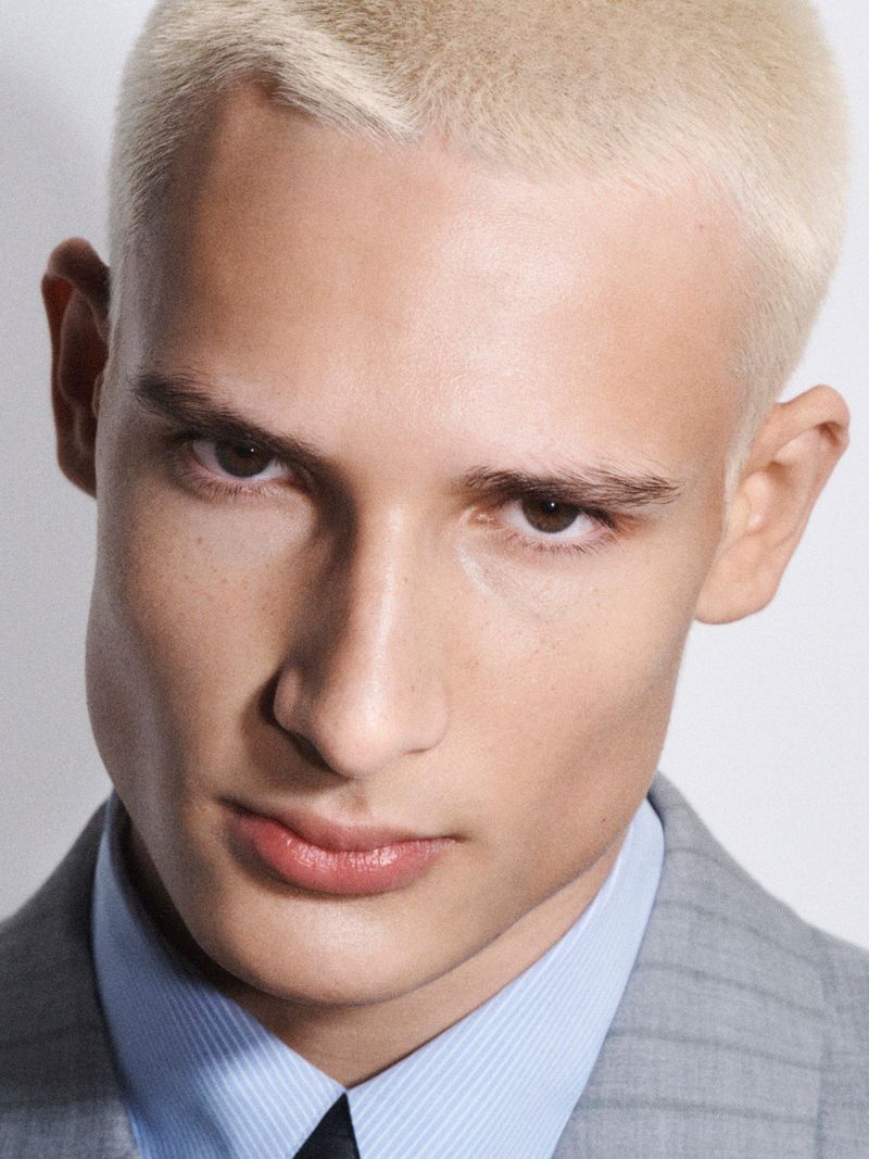 Close-up portrait of an adult male with bleached blonde hair wearing a grey pinstripe suit, a blue shirt, and a navy-blue tie in a photo studio.