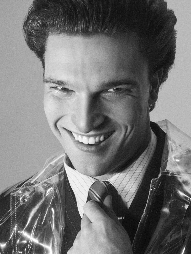 Monochrome portrait of a smiling adult male in a suit and transparent jacket, adjusting his tie against a plain background.