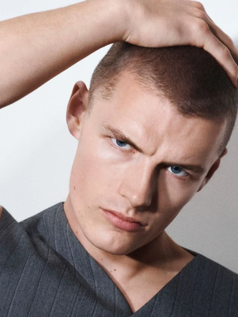 Close-up shot of a young adult man with a very short haircut and bright blue eyes, posing with his hand on top of his head.