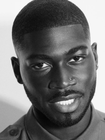 Close-up black and white portrait of a Black man with a fade haircut, groomed beard, and nose stud, smiling gently at the camera.