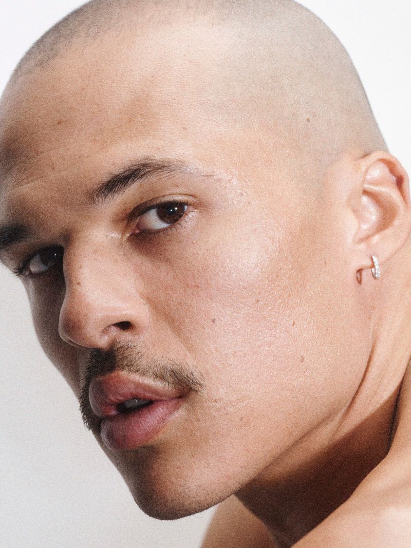 Close-up studio shot of an adult male with a shaved head and a mustache, wearing an earring against a plain white backdrop.