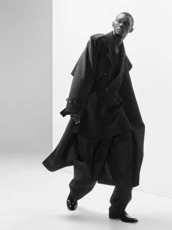 Model Fernando Cabral walks in an oversized black layered trench coat and wide trousers against a white studio background.