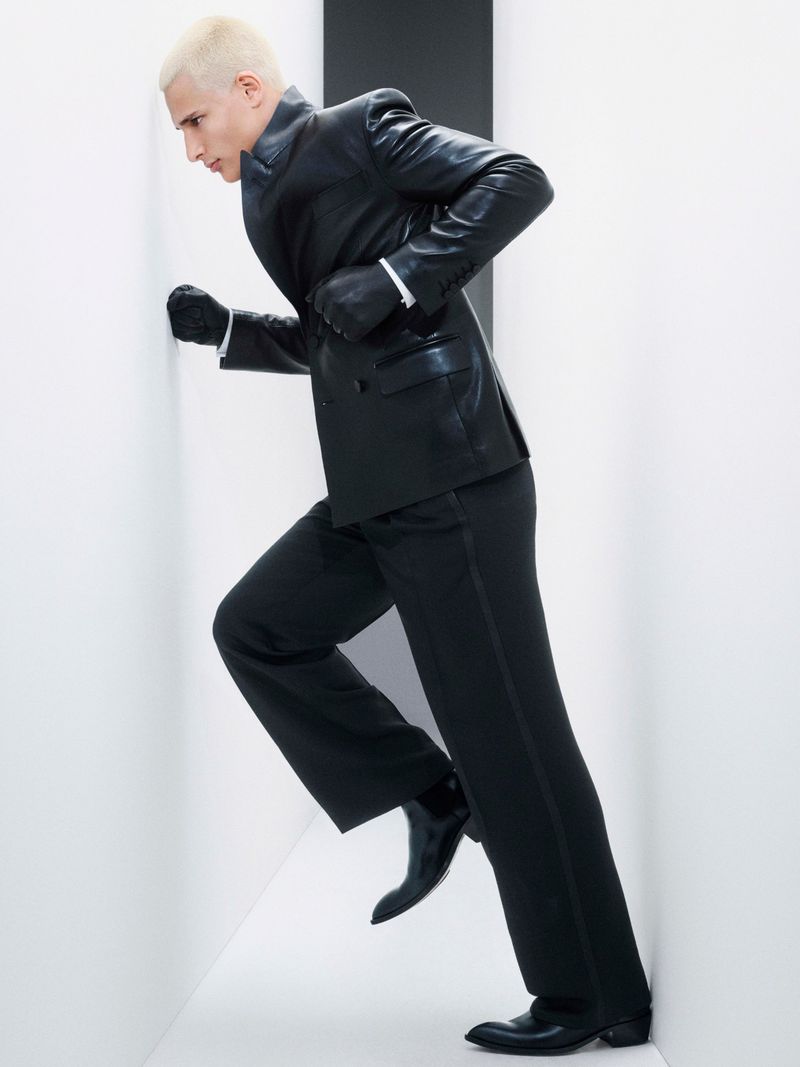 Fashion model in black suit strikes dynamic pose in narrow white space with contrasting black stripe, under bright, even lighting.
