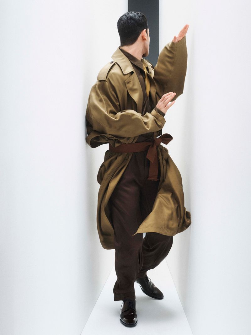 Man poses in a trench coat and brown trousers against a bright white minimalist hallway. He faces away from the camera.