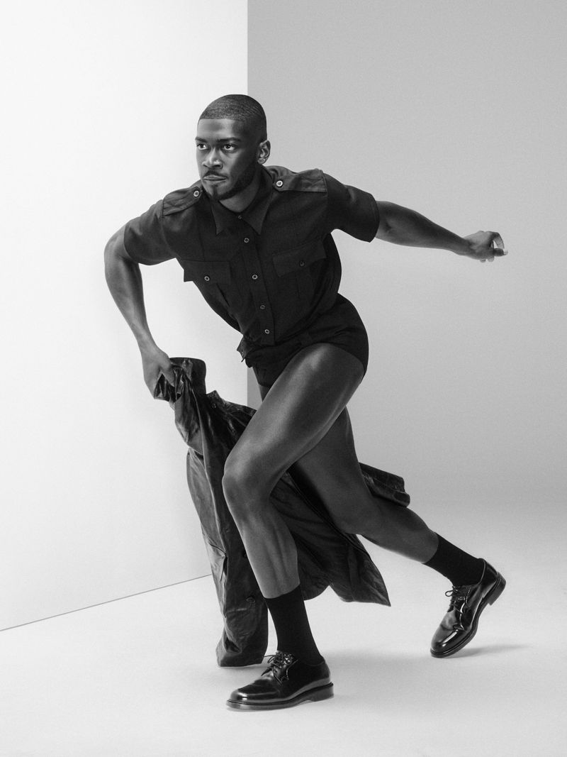A black and white studio shot features a Black man in a dark outfit, caught mid-stride, holding a coat, against a minimalist backdrop.