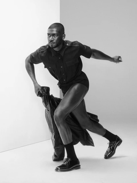 A black and white studio shot features a Black man in a dark outfit, caught mid-stride, holding a coat, against a minimalist backdrop.