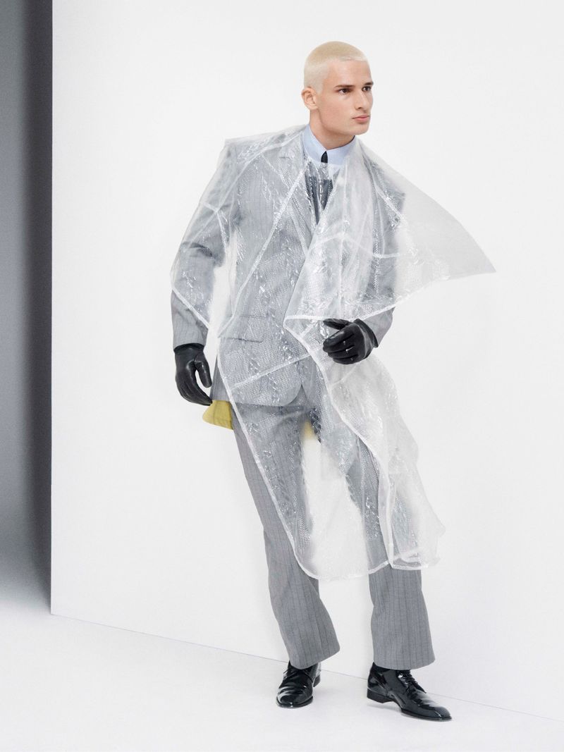 Model with platinum blonde hair wearing a grey pinstripe suit and transparent plastic overlay against a white studio background.