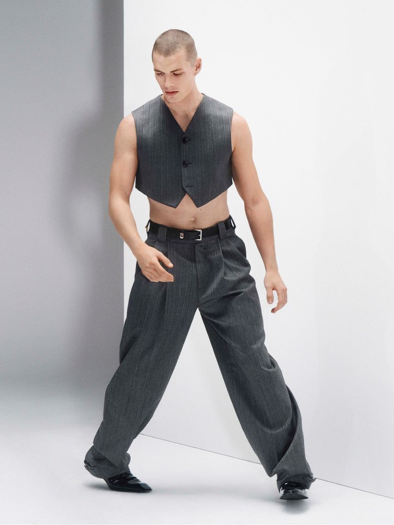 Young adult male poses in a gray pinstripe vest and wide-legged pants against a simple studio backdrop, conveying a modern fashion statement.