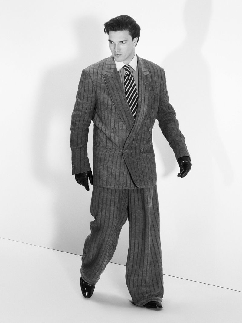 Man in a pinstripe suit walks in a brightly lit studio, wearing black gloves and black patent leather shoes against a white backdrop.