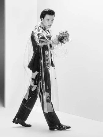 Monochrome fashion shot of a man in a suit and transparent coat, holding flowers, captured in a white studio with stylish lighting.