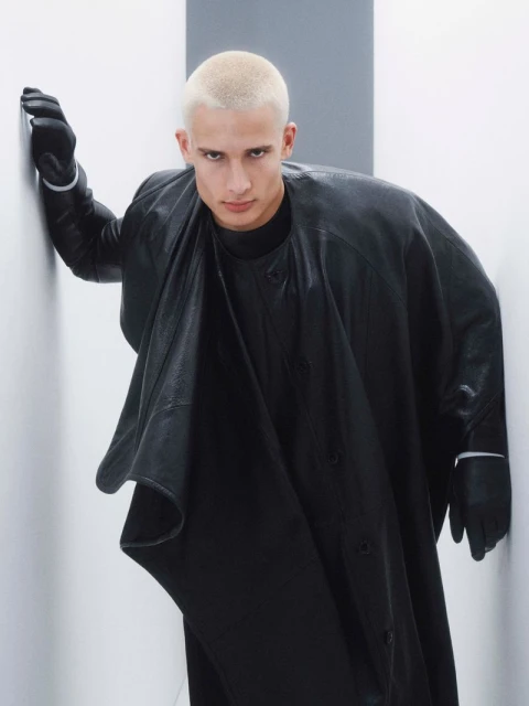 A young, blonde man with a shaved head stands indoors wearing a dark coat, gloves, and shirt against a gray-and-white wall in a studio shot.