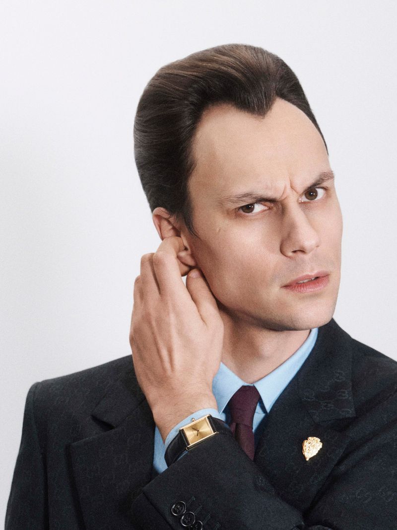 Close-up studio portrait of a man in a dark suit and light blue shirt with his hand near his ear, looking intensely at the camera.