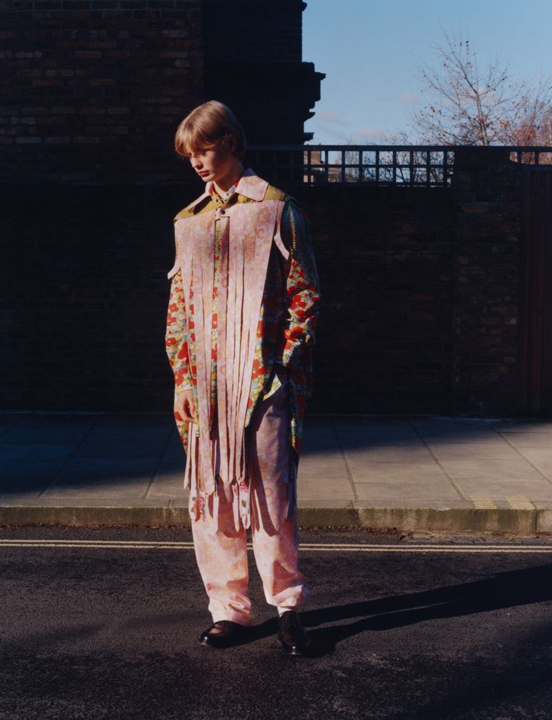 A person with blond hair stands outside on an asphalt street wearing patterned clothing and a vest with fringe in the daylight.
