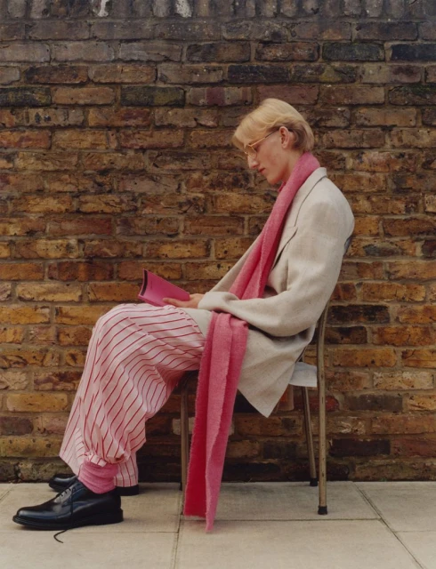 A person sitting on a chair outside of a brick building, reading from a bright pink book and wearing a pink scarf.