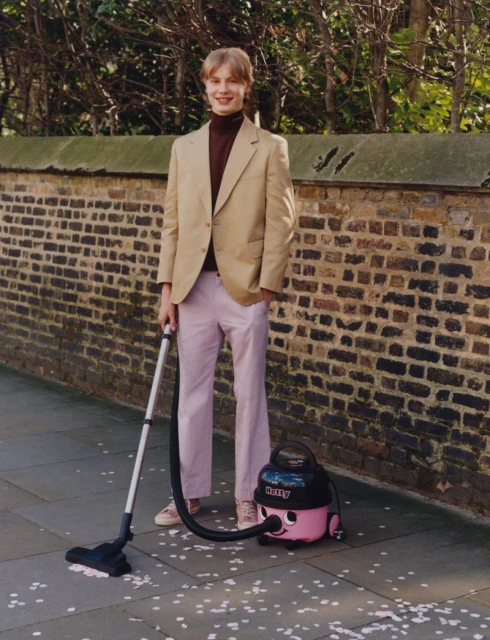 A young adult stands outside in stylish clothing next to a pink vacuum cleaner with a smiling face; brick wall and budding trees.