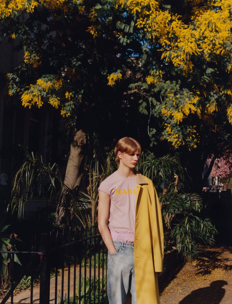 Young adult stands in front of a green tree and yellow flowers wearing a pink t-shirt, light blue jeans, and a yellow coat draped on their shoulder.