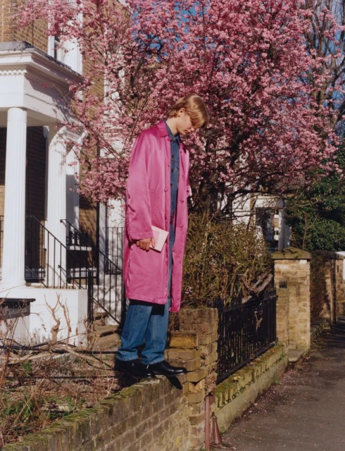 Stylish adult in a pink coat poses on a brick wall near a house with pink cherry blossom in background on a sunny day.