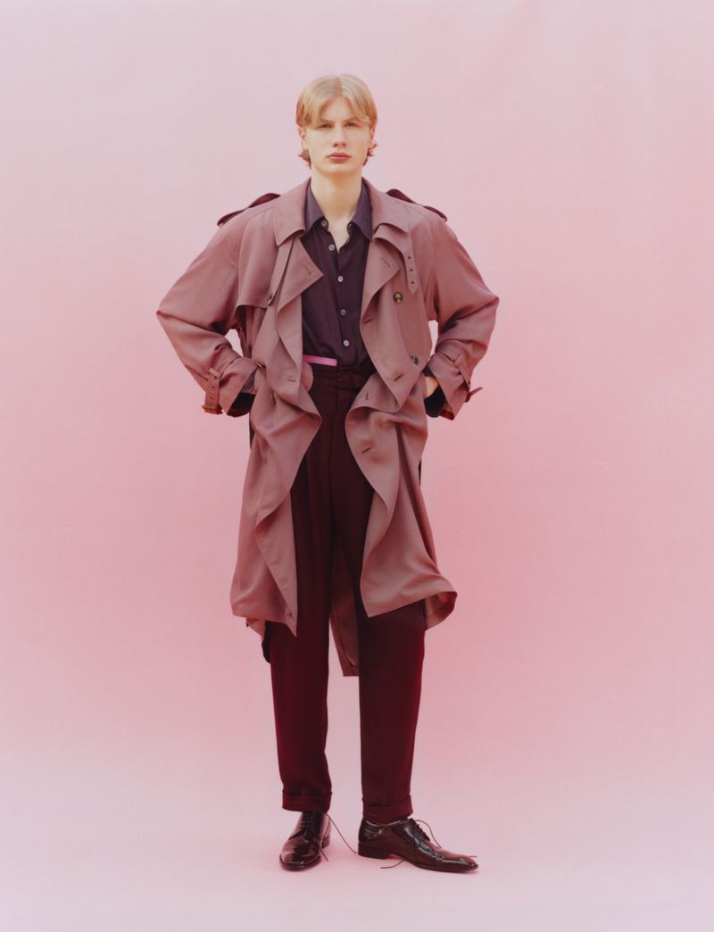 Model Orlando Straub standing in an oversized dusty rose trench coat and dark trousers against a pink studio backdrop.