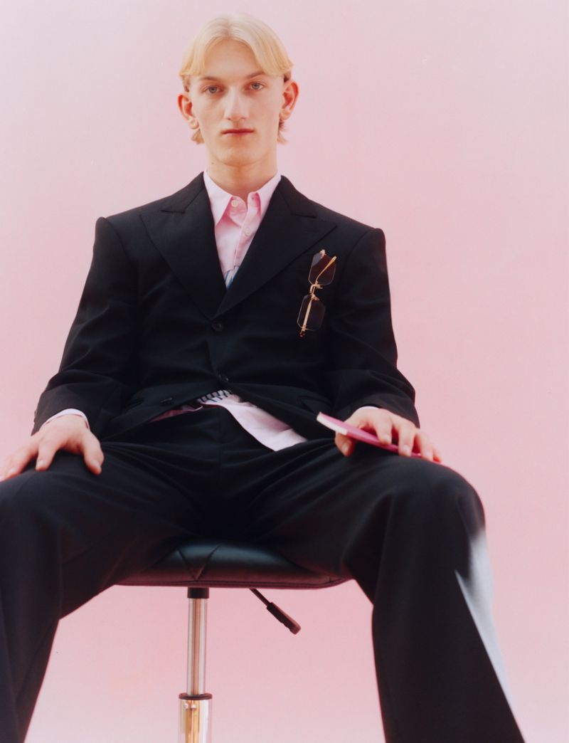 Young adult with blonde hair in a black suit and pink shirt sits in a rolling chair against a pink backdrop, holding a pink object.