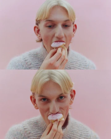 Close-up of a young adult with blond hair, wearing a sweater, eating a frosted donut against a pale pink background in a portrait-style composition.