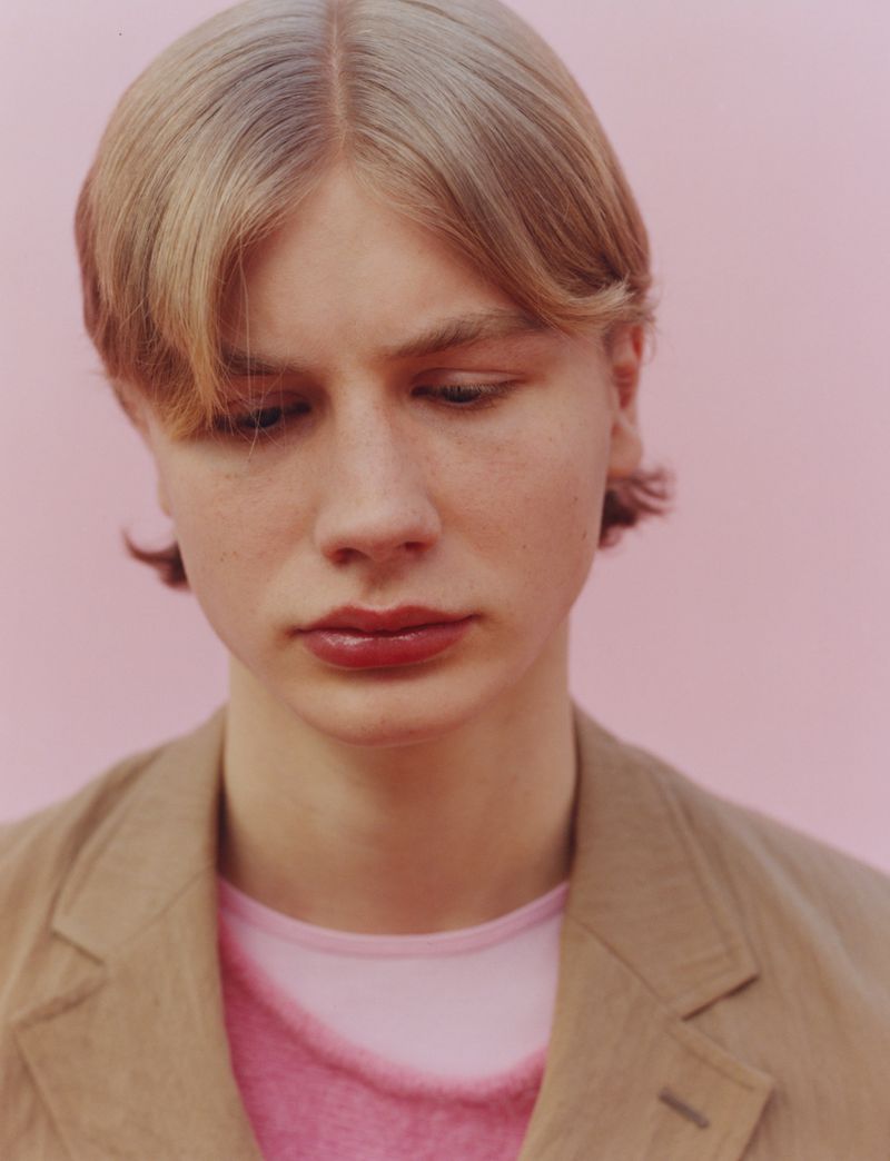Portrait of a fair-skinned person with blonde hair in a tan jacket over a pink shirt, standing against a pink background in a studio shot.